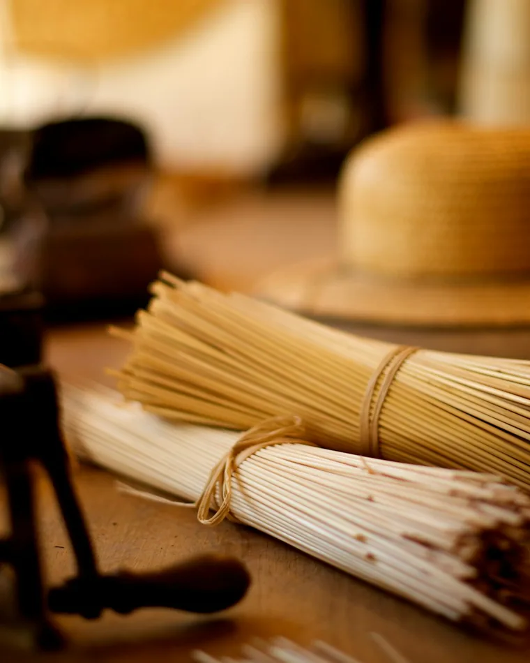 Mazos de paja natural atados con hilo sobre una mesa de taller, con un sombrero de paja claro y herramientas al fondo