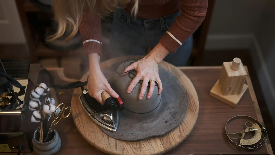 Planchado al vapor de una campana de fieltro gris sobre una horma de madera, con tijeras, cinta métrica y utensilios de sombrerería alrededor